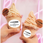 Two soft serve ice cream cones with branded mini wafers held by hands, branded 'The Madison Club' on a pink background.